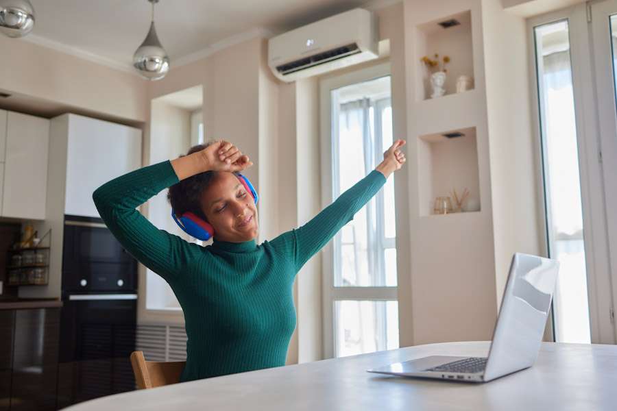 Overjoyed millennial girl wearing headphones have fun moving listening to music relax in living room, happy young woman in earphones dance enjoy leisure weekend at home, stress free concept Wohnraum mit Split-Klimaanlage über dem Fenster; Person mit Laptop am Tisch in klimatisiertem Raum in moderner Wohnung.